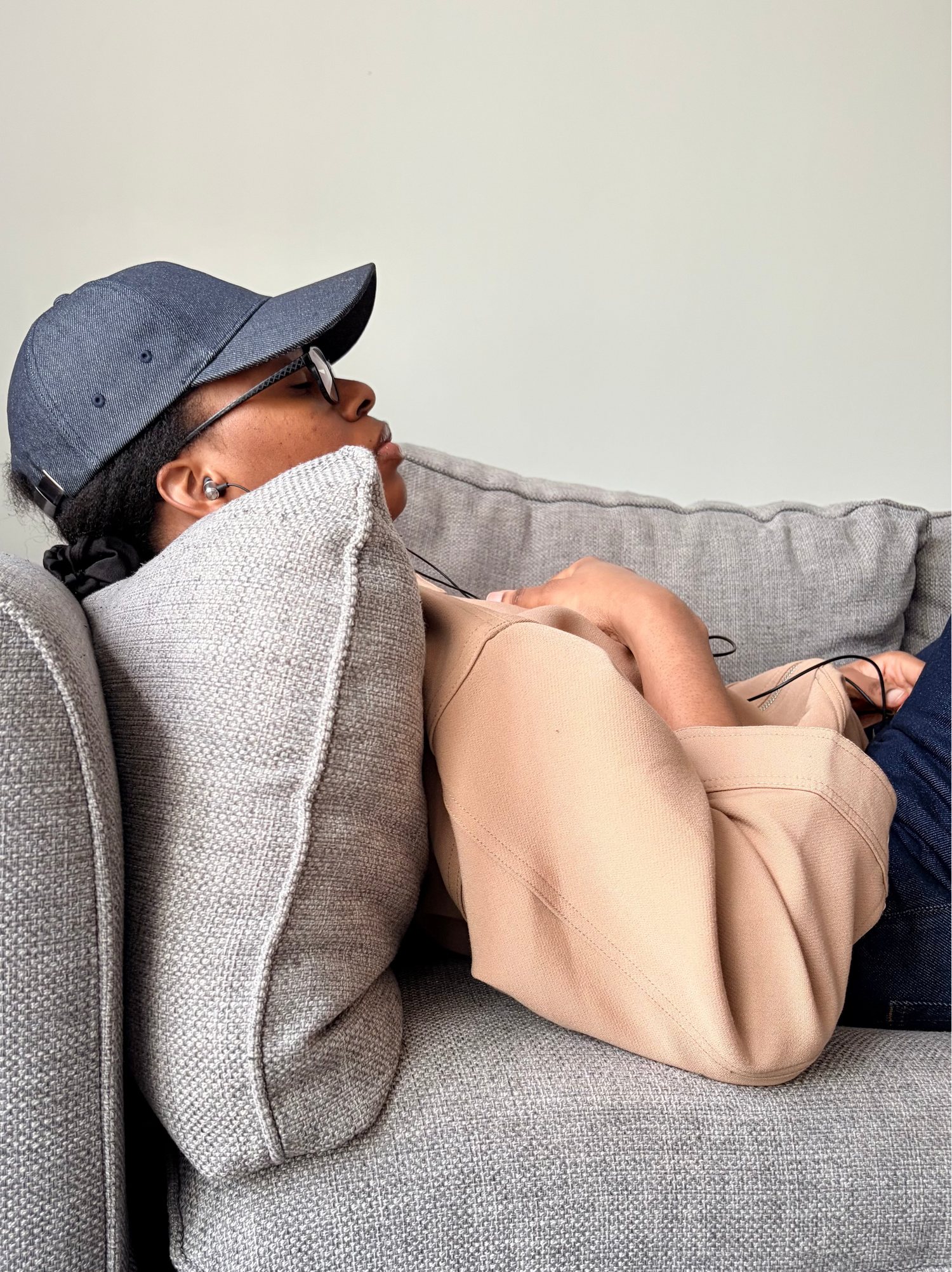 Person sitting on a gray couch with a pillow, wearing a blue cap and beige jacket.