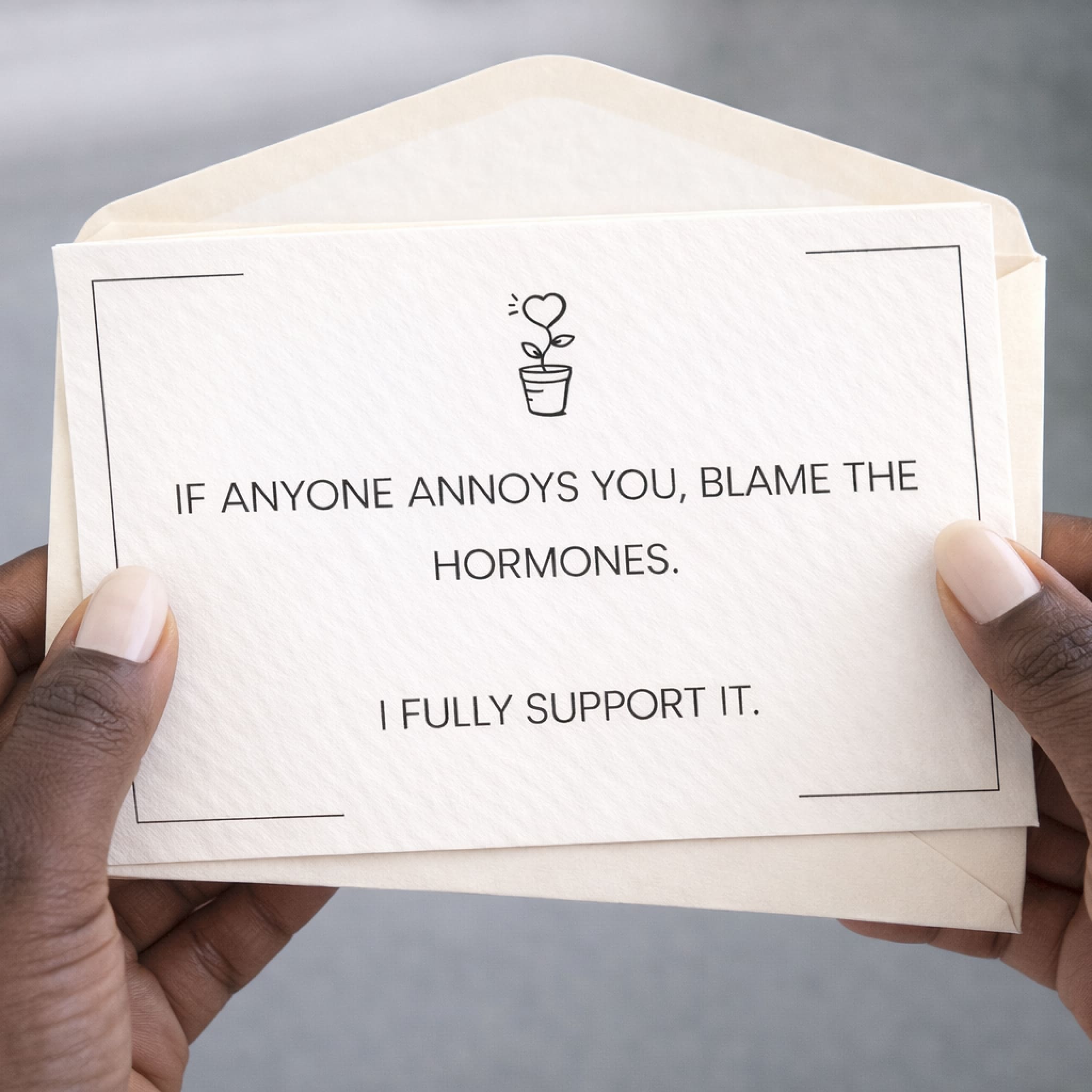 Envelope with humorous message about blaming hormones held by hands against a neutral background