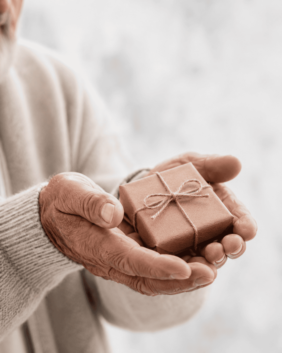 Close-up of elderly hands holding a small brown gift box with a bow against a blurred background.